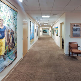 A hallway gallery with various artworks displayed on both sides, including a large painting of a woman with a hippo in the foreground. The hallway extends with additional framed pieces, creating a bright, inviting exhibition space.