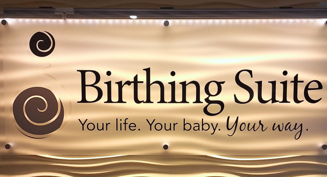 A softly lit sign for "Birthing Suite" featuring elegant text and swirled decorative accents, with the tagline "Your life. Your baby. Your way." in a welcoming font