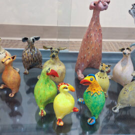 A collection of vibrant ceramic animals, including whimsical birds and animal figures, arranged on two shelves in a glass display case.