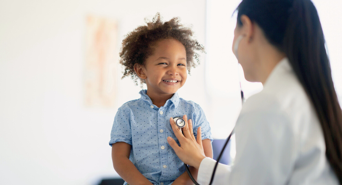 A smiling young child in a blue shirt having their heartbeat checked by a healthcare provider with a stethoscope in a bright, clean medical office.