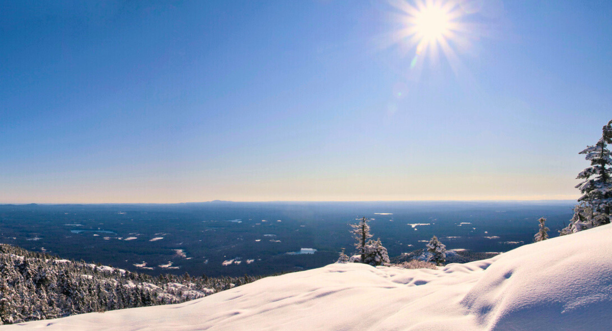 A serene winter landscape with a snow-covered slope, evergreen trees dusted with snow, and a bright sun shining in a clear blue sky, overlooking a vast expanse of forested terrain and distant lakes