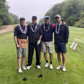 Four golfers dressed in themed attire, including leis and a grass skirt, smiling on the green at Monadnock Community Hospital's Fall Foliage Golf Classic