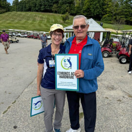 Two individuals smiling and holding a sponsor sign for "Edmunds Ace Hardware" at Monadnock Community Hospital's Fall Foliage Golf Classic, with a backdrop of golf carts and greenery