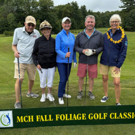 Five golfers standing on the green at Monadnock Community Hospital's Fall Foliage Golf Classic, posing behind a banner that reads "MCH Fall Foliage Golf Classic," with trees and fairways in the background