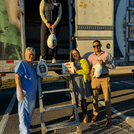 Four smiling individuals pose for a photo near a truck, with one person standing in the truck doorway holding a bagged turkey. The others stand on the ground by a ladder, holding bagged turkeys and a sign. The setting appears to be outdoors in bright sunlight