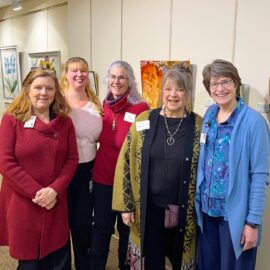 Five smiling women pose together in front of displayed artwork at Monadnock Community Hospital’s Healing Arts Gallery during the Birds of a Feather opening.