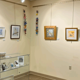 Framed bird-themed artwork and origami cranes are displayed on cream-colored walls in a corner of the Healing Arts Gallery, with a glass case of small bird sculptures and prints below.