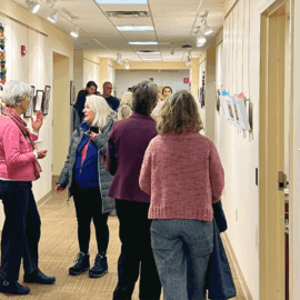 Guests gather and chat in the hallway of the Healing Arts Gallery during the Birds of a Feather opening reception, with framed artwork and colorful paper cranes lining the walls.