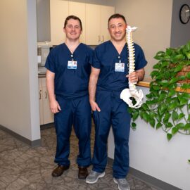 Dr. Vache Hambardzumyan and Tom Quinn standing side by side in medical scrubs, smiling in a clinic setting. Dr. Hambardzumyan is holding a model of a spine, symbolizing their dedication to orthopedic care