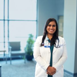 Dr. Mandalika, an internal medicine physician, standing confidently in a bright and welcoming hospital setting, wearing a white coat and stethoscope, with a friendly smile. The background features seating and a plant, creating a professional yet approachable atmosphere