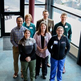 Seven Monadnock Community Hospital employees standing together in a bright space with floor-to-ceiling windows, wearing professional attire and smiling warmly, showcasing their teamwork and community focus