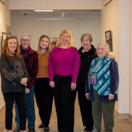 Six members of the Healing Arts Gallery Committee standing together in an art-filled hallway, smiling warmly and representing their dedication to bringing creativity and inspiration to Monadnock Community Hospital