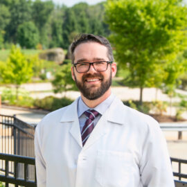 Dr Spencer in a white lab coat, wearing glasses and a purple tie, smiling outdoors with trees and a landscaped background