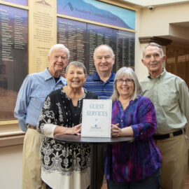 Five volunteer greeters at Monadnock Community Hospital standing in the lobby by the guest services sign, smiling warmly to highlight their commitment to welcoming patients and visitors