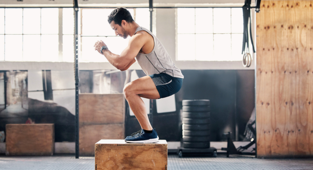 Man performing a box jump in a gym, wearing a grey tank top and black shorts. He is landing on a wooden box with knees bent, engaging his leg muscles in a controlled movement]