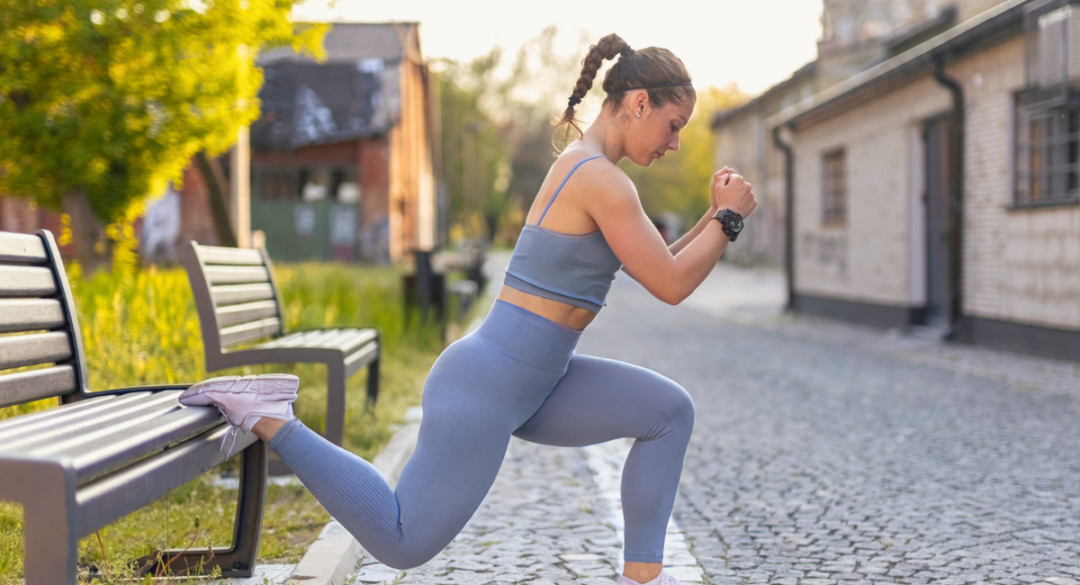 Woman performing a Bulgarian split squat outdoors using a park bench for support. She is dressed in gray workout gear, concentrating on her form as she strengthens her lower body