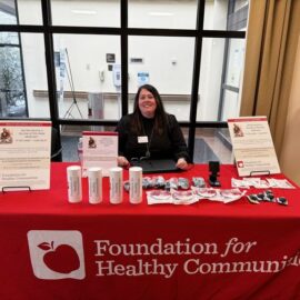 A representative from the Foundation for Healthy Communities sitting behind a promotional table covered with branded materials and informational brochures at a health fair in partnership with Monadnock Community Hospital