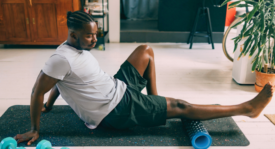 Man using a foam roller on his lower leg while sitting on a yoga mat indoors. He is wearing a white t-shirt and black shorts, focusing on relieving muscle tension