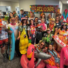 Staff from Monadnock Orthopaedic Associates dressed in tropical beach-themed costumes, including leis, inflatable pool toys, and Hawaiian shirts, posing in a festive setting with a sign reading 'Aloha.'