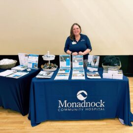 Monadnock Community Hospital's Human Resources department booth at a health fair, featuring brochures, promotional items, and a staff member standing behind the table dressed in a branded blue shirt