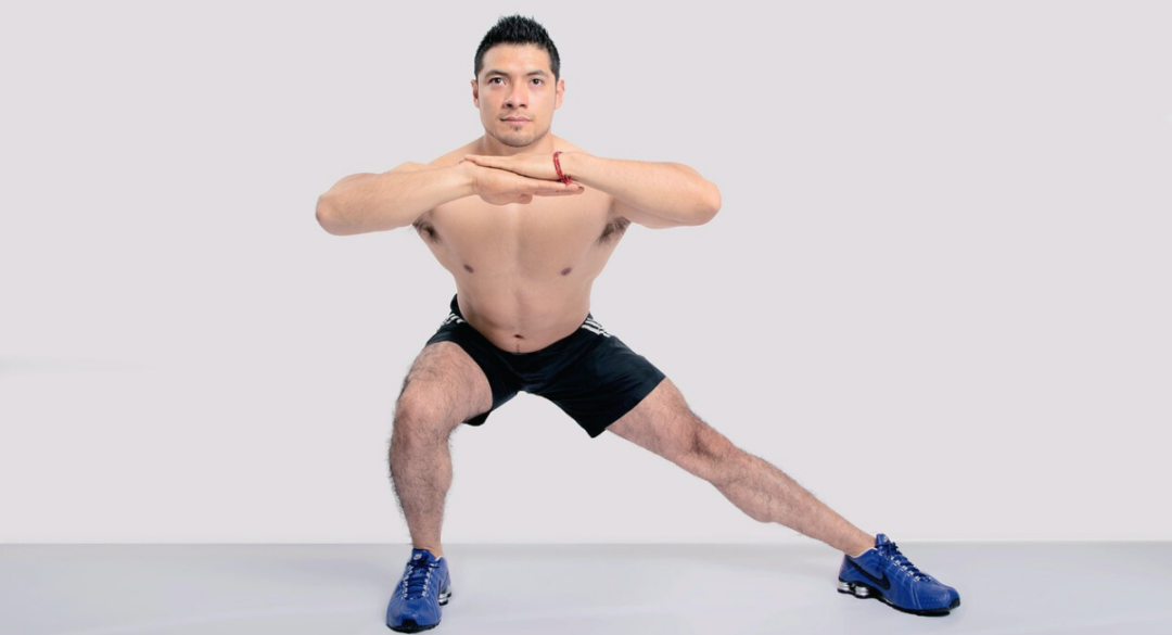 Man performing a lateral lunge, wearing black shorts and blue sneakers. He is focusing on stretching and strengthening his inner thighs and glutes in a fitness pose