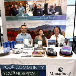 Three Monadnock Community Hospital representatives standing behind a recruitment booth. The booth features promotional materials, water bottles, and a backdrop highlighting community, hospital, and career opportunities with scenic imagery of Mount Monadnock