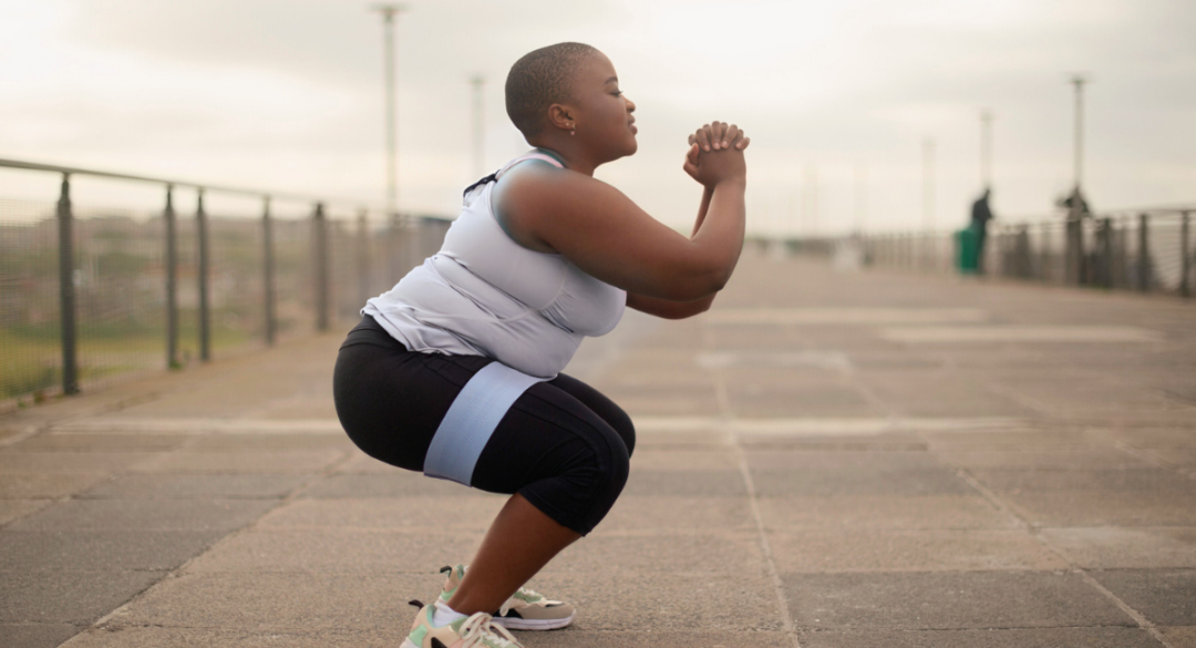 Woman performing a squat outdoors on a concrete pathway, wearing a gray tank top, black leggings, and resistance band around her thighs. She is focused on her form, with hands clasped in front, engaging her legs and core muscles.