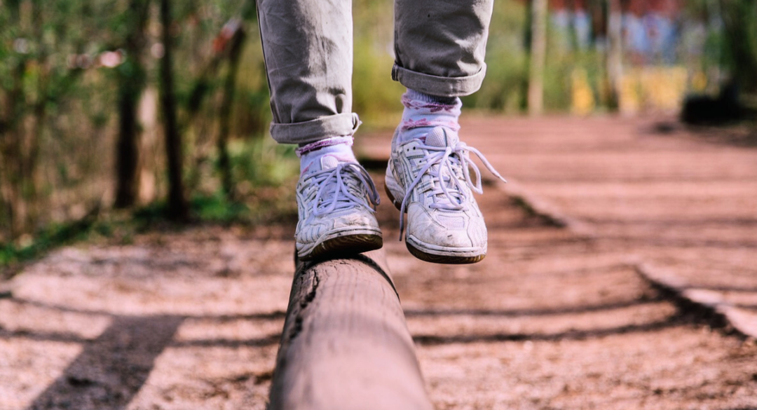Person balancing on a wooden beam with one foot, wearing casual pants and white sneakers. The scene takes place on a wooded trail, focusing on stability and balance