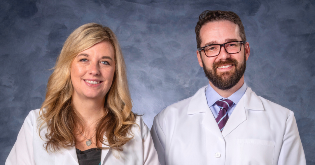 Two healthcare providers wearing white lab coats stand side by side against a blue studio backdrop, smiling at the camera. One has shoulder-length blonde hair and wears a teal top, and the other has short dark hair, glasses, a beard, and a striped tie.