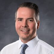 A smiling man with dark, neatly styled hair wearing a white dress shirt and a light blue patterned tie, posed in front of a solid dark gray background.