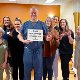 A group of healthcare professionals at Monadnock Community Hospital stands together in a clinical setting, smiling and holding up peace signs. In the center, a man in navy blue scrubs holds a sign that reads, "2,000 PROCEDURES COMPLETED – Monadnock Community Hospital." The group includes individuals in scrubs, casual clothing, and business attire, all celebrating the milestone. The background features an orange accent wall, medical curtains, and hospital equipment.