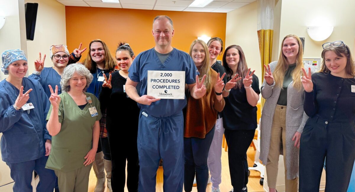 A group of healthcare professionals at Monadnock Community Hospital stands together in a clinical setting, smiling and holding up peace signs. In the center, a man in navy blue scrubs holds a sign that reads, "2,000 PROCEDURES COMPLETED – Monadnock Community Hospital." The group includes individuals in scrubs, casual clothing, and business attire, all celebrating the milestone. The background features an orange accent wall, medical curtains, and hospital equipment.