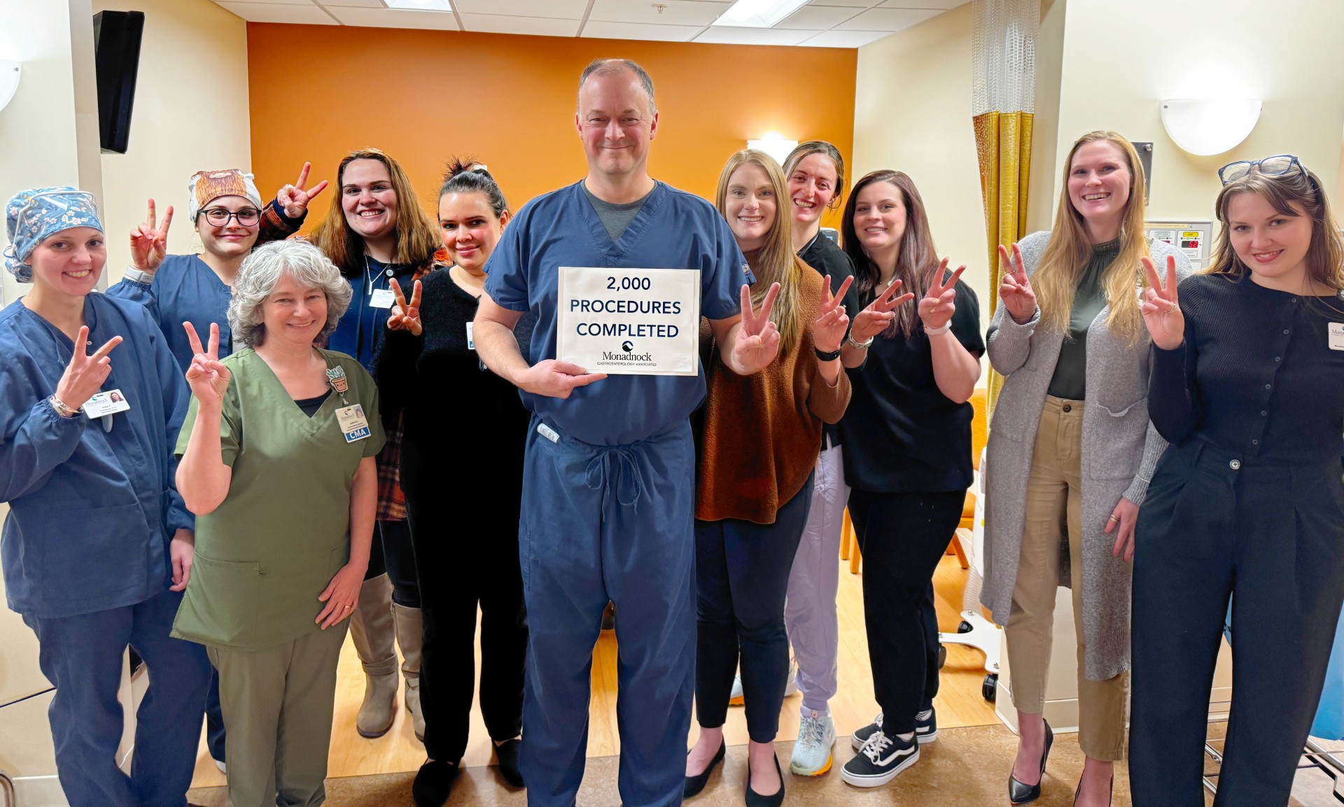 A group of healthcare professionals at Monadnock Community Hospital stands together in a clinical setting, smiling and holding up peace signs. In the center, a man in navy blue scrubs holds a sign that reads, "2,000 PROCEDURES COMPLETED – Monadnock Community Hospital." The group includes individuals in scrubs, casual clothing, and business attire, all celebrating the milestone. The background features an orange accent wall, medical curtains, and hospital equipment.