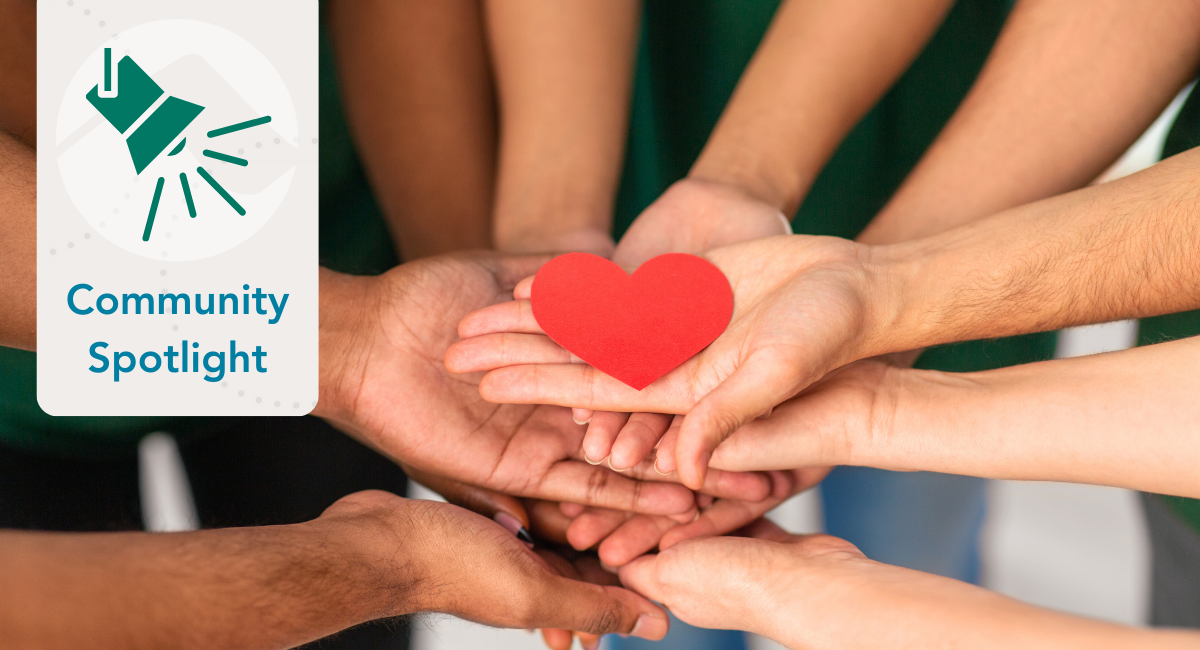 Community Spotlight banner featuring a green spotlight icon and the text 'Community Spotlight' on the left. On the right, several hands of diverse individuals are cupped together, holding a red heart, symbolizing unity and compassion. The background is blurred, creating a warm and inviting atmosphere