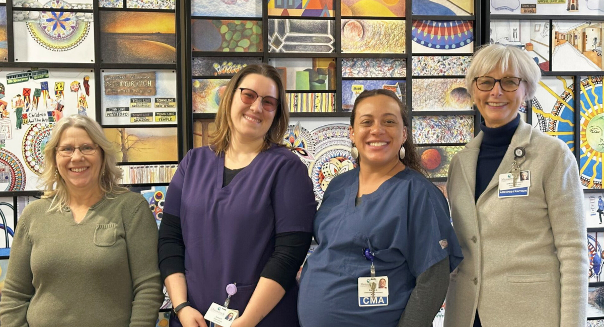 Four women standing in front of a colorful mosaic artwork. From left to right: Sherry Olson, Deiana Gaudette, Brianna Blake, and Cynthia McGuire. All are smiling and wearing professional attire.