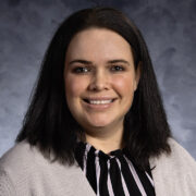 A woman with shoulder-length dark hair and light skin stands in a professional office setting, smiling at the camera. She is wearing a black-and-white striped blouse under a light gray cardigan. The background features a brick hallway and a beige wall with a framed painting of a sailboat.