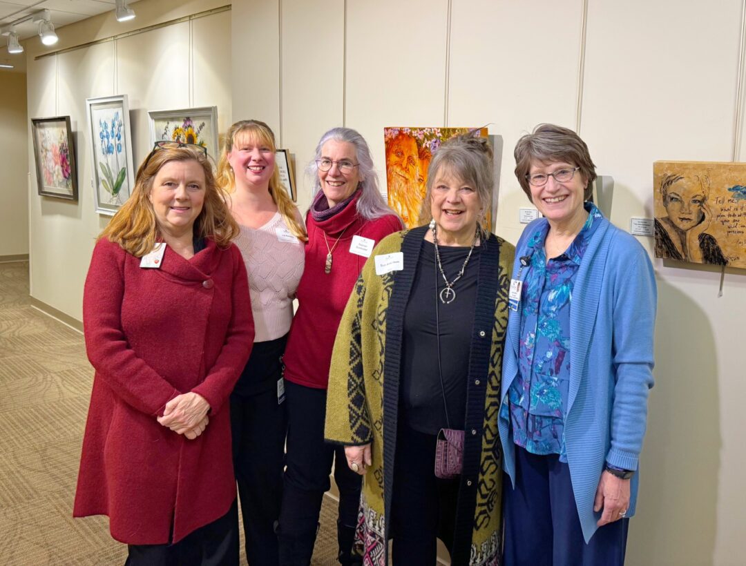 Five women stand together in front of artwork at the Healing Arts Gallery opening. From left to right: Katharyn Ernst, Liz Kenney, Sooson Dunholter, artist Sue Ann Hum, and Vice President of Philanthropy & Community Relations Laura Gingras. The gallery walls behind them feature a variety of framed paintings and mixed media pieces