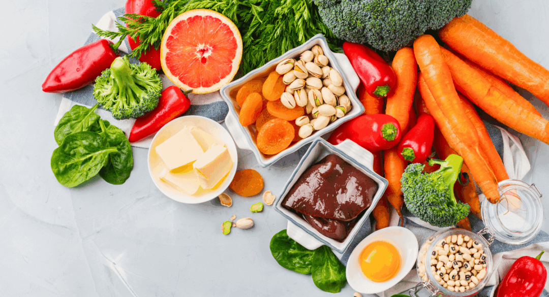 A colorful assortment of nutrient-rich foods, including carrots, broccoli, red peppers, spinach, grapefruit, dried apricots, pistachios, eggs, butter, and a dish of liver. The image highlights a balanced diet with a variety of vitamins and minerals essential for overall health.