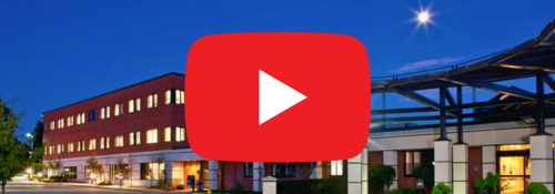 Exterior view of Monadnock Community Hospital at night, illuminated under a starry sky. A large YouTube play button overlay is centered, indicating a link to video content