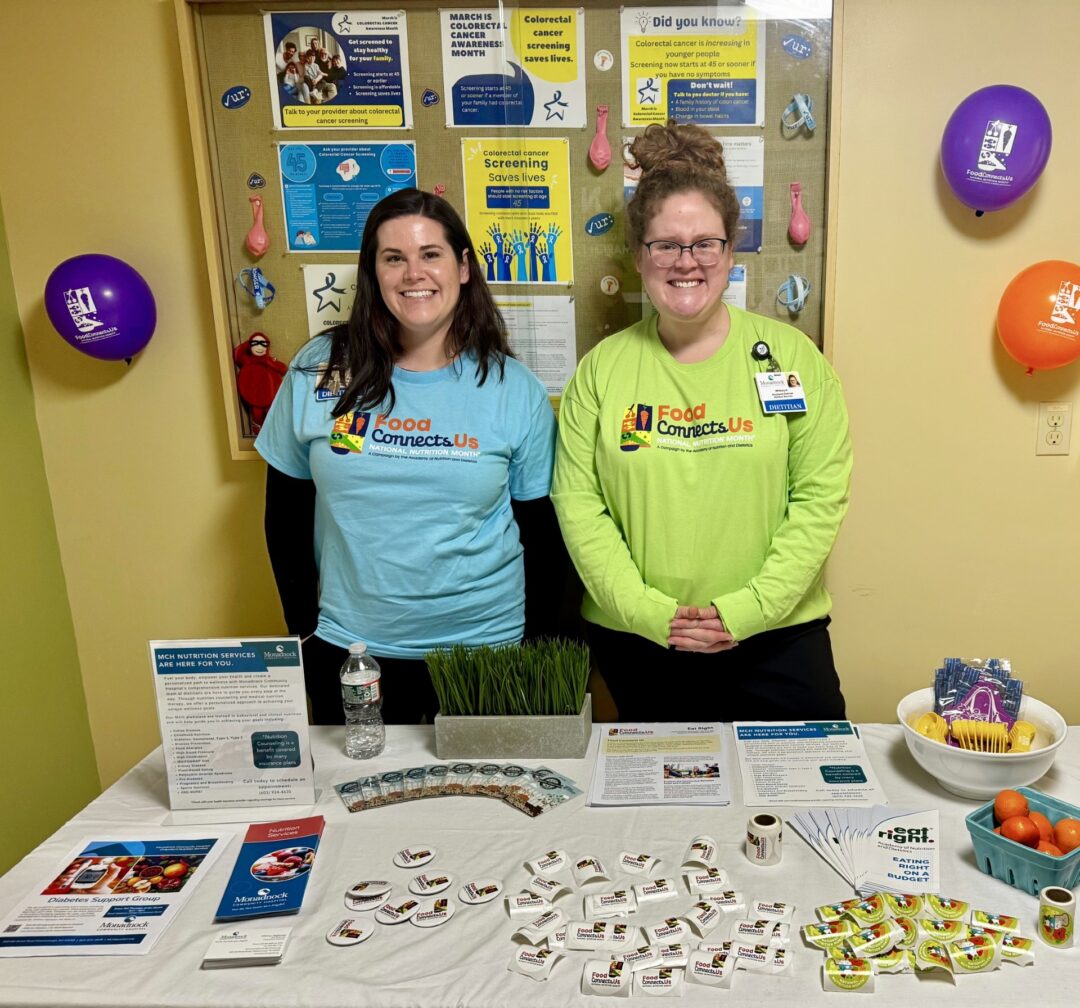 Kaitlyn McCarthy and Whitney Hightower from Monadnock Community Hospital’s Nutrition Services Department stand smiling behind a colorful nutrition education table for National Nutrition Month. The display includes wellness handouts, fruits, buttons, and brochures promoting balanced eating, with posters about colorectal cancer awareness in the background.