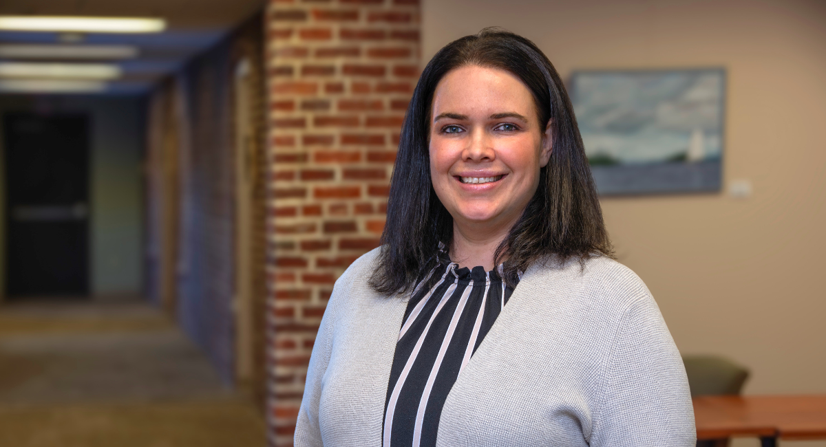 A woman with shoulder-length dark hair and light skin stands in a professional office setting, smiling at the camera. She is wearing a black-and-white striped blouse under a light gray cardigan. The background features a brick hallway and a beige wall with a framed painting of a sailboat.