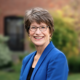 A smiling woman with short gray hair and glasses stands outdoors in front of a brick building. She is wearing a royal blue blazer over a black top and has her hands gently clasped in front of her. The background features green grass, a curved pathway, and autumnal foliage.