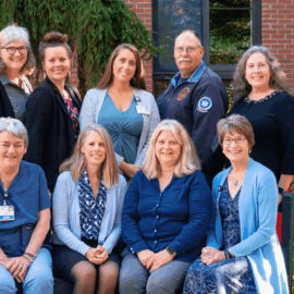 A group of eleven smiling adults poses outside a brick building, with greenery in the background. Some are seated in front, while others stand behind them. They represent a mix of healthcare, emergency services, and community professionals, reflecting collaboration in community health initiatives.