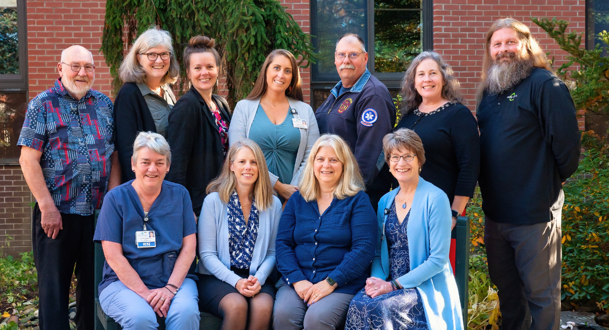 A group of eleven smiling adults poses outside a brick building, with greenery in the background. Some are seated in front, while others stand behind them. They represent a mix of healthcare, emergency services, and community professionals, reflecting collaboration in community health initiatives.
