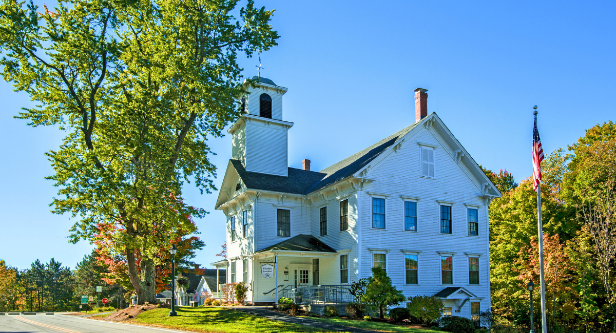 A white wooden town hall with a prominent bell tower and ramp-accessible entrance, surrounded by trees just beginning to turn with autumn colors. An American flag stands in the foreground.