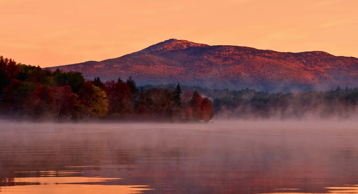 Mount Monadnock glows in warm hues at sunrise above a mist-covered lake, with reflections of fall-colored trees and shoreline homes in the still water.