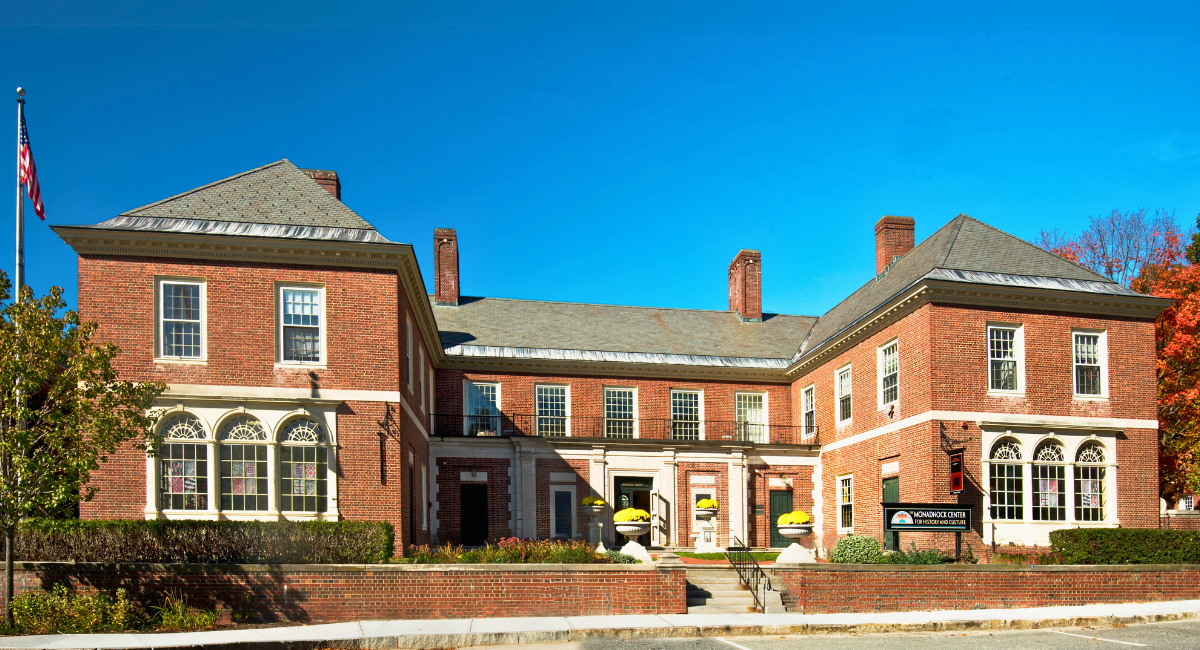 A stately brick building with white trim and arched windows, flying the American flag on a clear blue day. The sign reads "Monadnock Center for History and Culture," and yellow potted mums decorate the front steps.