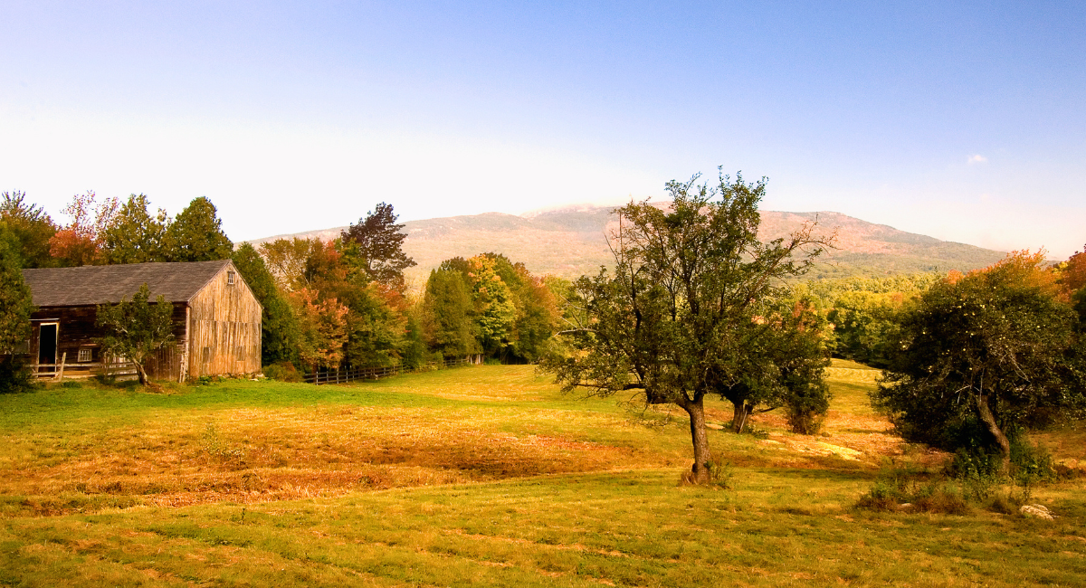 A rustic wooden barn stands at the edge of an open grassy field with scattered trees in early fall foliage, framed by a distant view of Mount Monadnock beneath a soft blue sky.