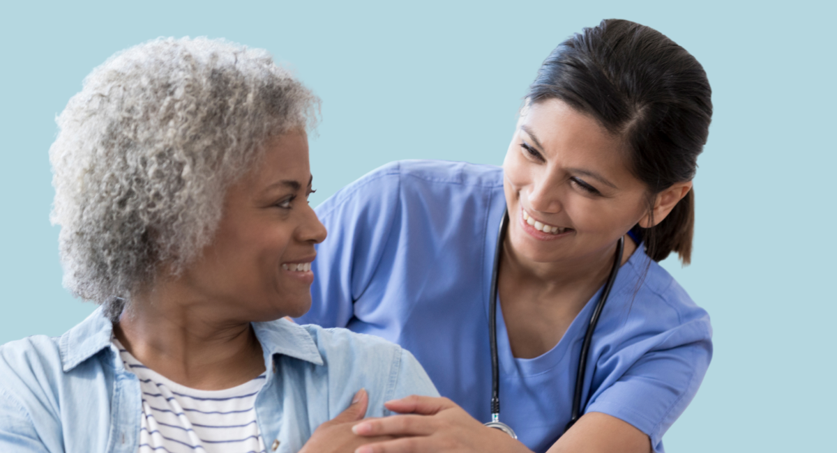 Smiling healthcare provider in blue scrubs with a stethoscope warmly interacts with an older adult patient with curly gray hair, both looking at each other with friendly expressions against a light blue background.