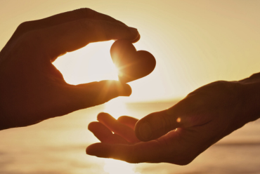 Two hands holding a heart-shaped rock, with the sun setting in the background, symbolizing compassion and connection.